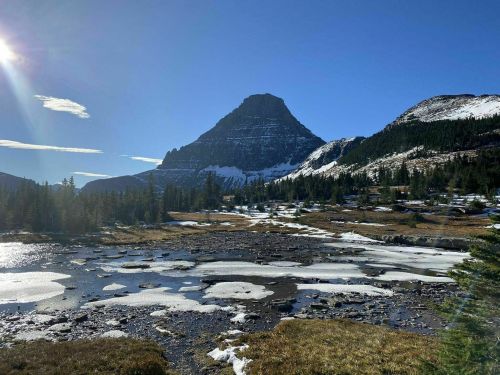 A scenic mountain landscape with a peak, clear blue sky, and patches of snow near a reflective pond.