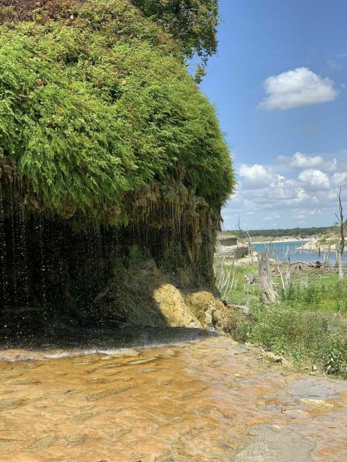 A lush green cliff with cascading water, overlooking a serene lake under a blue sky with scattered clouds.