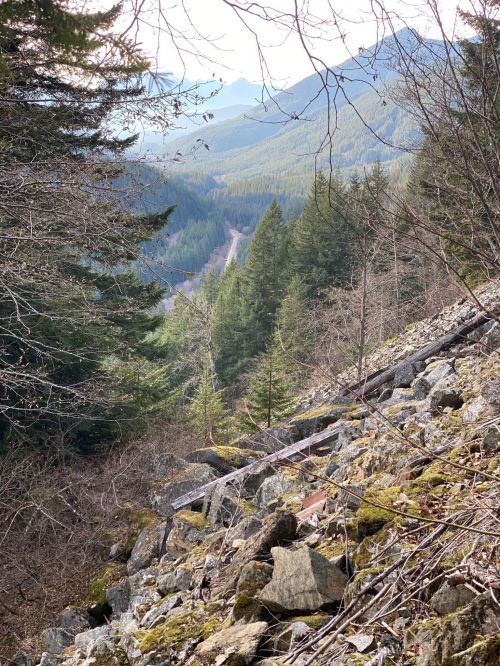 A rocky slope leads down to a valley surrounded by lush green mountains under a clear sky.