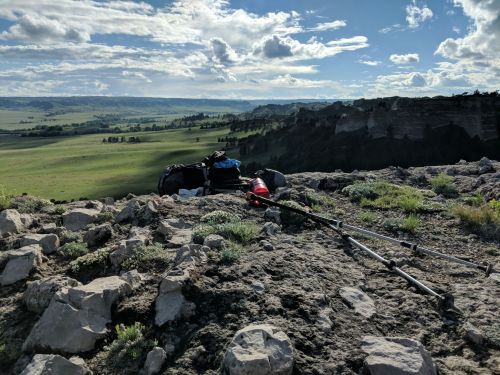 A rocky cliff overlooks a green valley under a blue sky with scattered clouds, with hiking gear in the foreground.