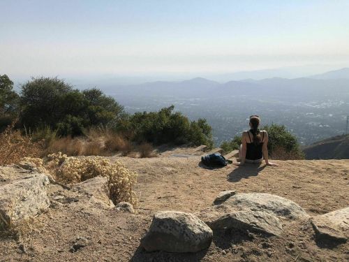 A person sits on a rocky hillside, gazing at a distant valley and mountains under a clear sky.