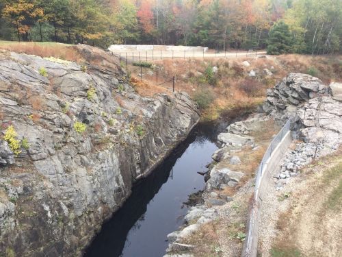 A rocky landscape with a narrow waterway, surrounded by autumn foliage and a fenced area in the background.