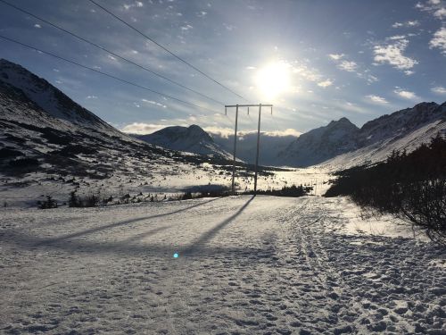 A snowy landscape with mountains, power lines, and a bright sun shining in a clear blue sky. Shadows stretch across the snow.
