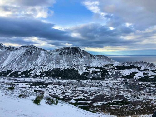 Snow-covered mountains under a cloudy sky, with a view of a valley and the ocean in the distance.