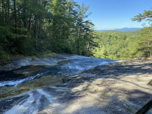 A serene landscape featuring a flowing stream over smooth rocks, surrounded by lush green trees and distant mountains.