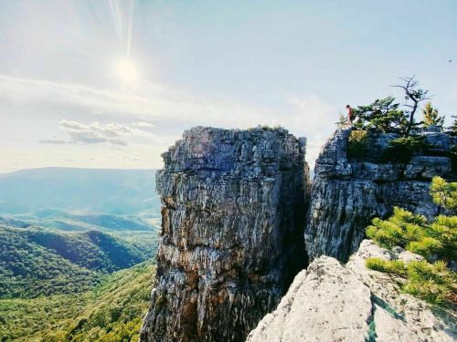 A person stands on a rocky cliff overlooking a lush green valley under a bright sun and blue sky.