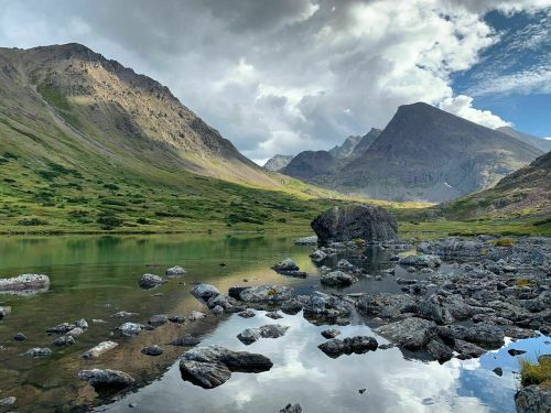 A serene mountain landscape with a calm lake, rocky shore, and dramatic clouds overhead.