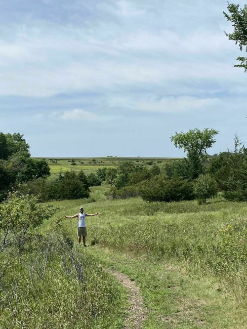 A person stands with arms outstretched in a grassy field, surrounded by trees and a wide, open landscape under a blue sky.