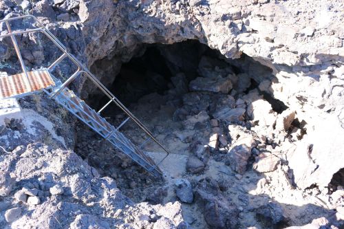 A metal staircase leads down into a rocky cave entrance surrounded by rough terrain.