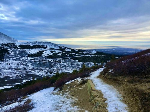 A winding path through a snowy landscape, with mountains and a cloudy sky in the background.
