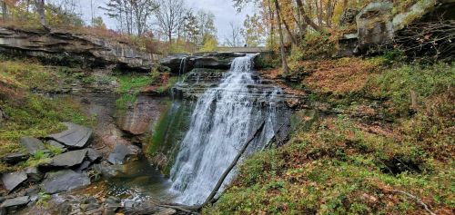 A serene waterfall cascades over rocky cliffs, surrounded by autumn foliage and a tranquil forest setting.