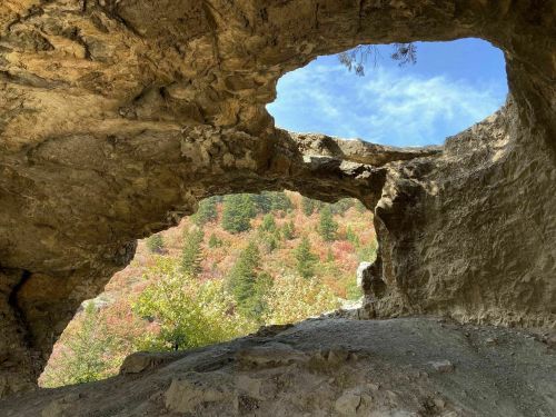 A rocky cave opening frames a view of colorful autumn trees and a blue sky.
