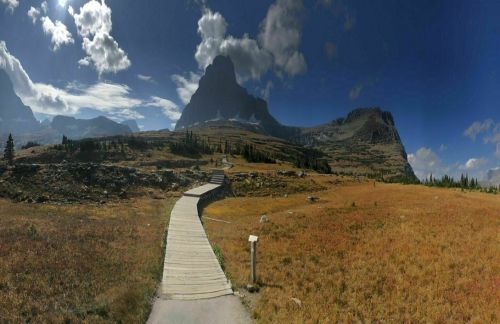 A wooden pathway leads through a grassy landscape towards a majestic mountain under a partly cloudy sky.