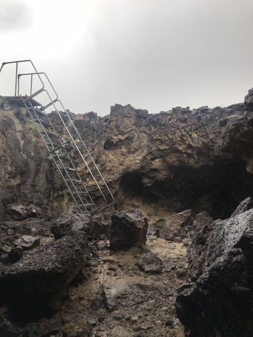 A rocky cave entrance with a metal ladder leading up to the surface, surrounded by dark volcanic rock.
