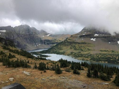A scenic view of mountains and a lake, partially obscured by clouds, with green trees in the foreground.