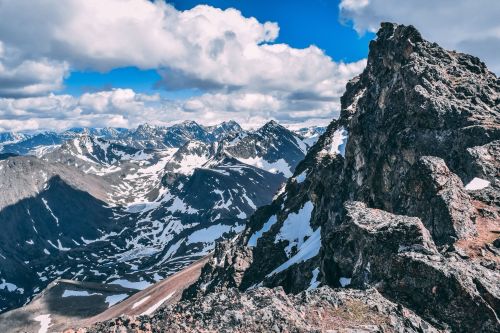 A panoramic view of rugged mountains with snow-capped peaks under a blue sky with fluffy clouds.