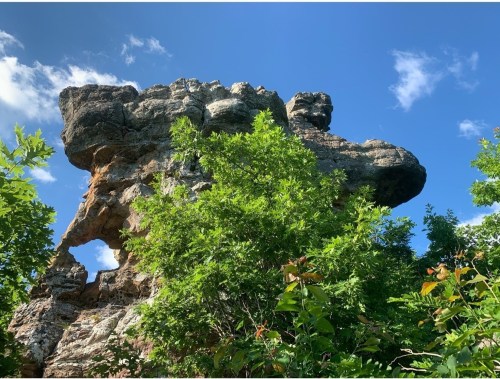 A large rock formation with a hole, surrounded by lush green foliage under a blue sky with scattered clouds.