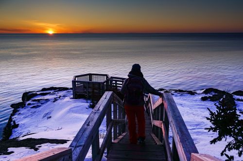 A person walks down wooden stairs towards a calm lake at sunset, with snow-covered rocks and a clear sky.