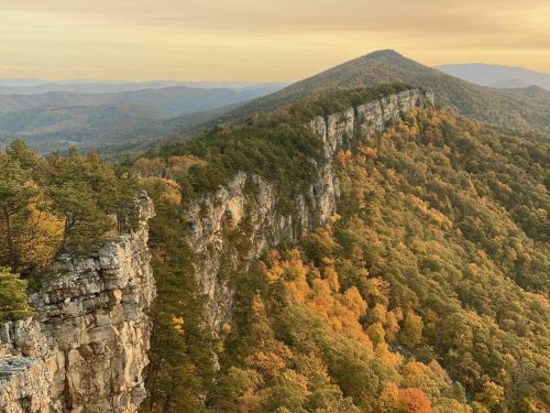 A scenic view of a rocky cliff surrounded by vibrant autumn foliage and distant mountains under a golden sky.