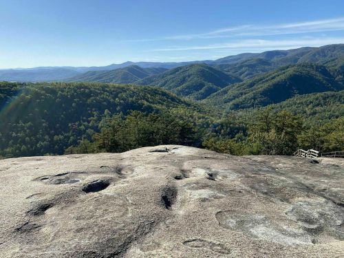 A panoramic view of lush green mountains under a clear blue sky, seen from a rocky outcrop.