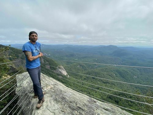 A person stands on a rocky ledge overlooking a lush green valley under a cloudy sky.