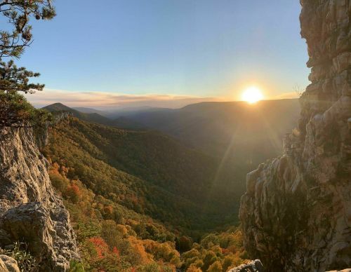 Sunset over a colorful valley, framed by rocky cliffs and trees, with warm light illuminating the landscape.
