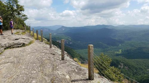 A rocky overlook with wooden posts and a scenic view of mountains and valleys under a partly cloudy sky.