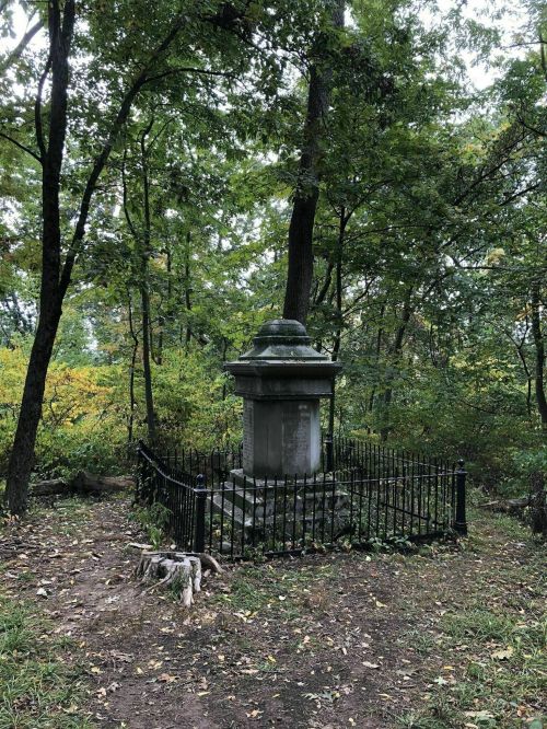 A weathered stone monument surrounded by trees, enclosed by a black iron fence in a wooded area.