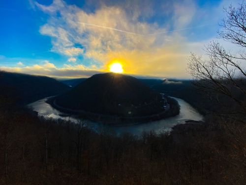 Sunrise over a winding river surrounded by mountains, with a colorful sky and bare trees in the foreground.