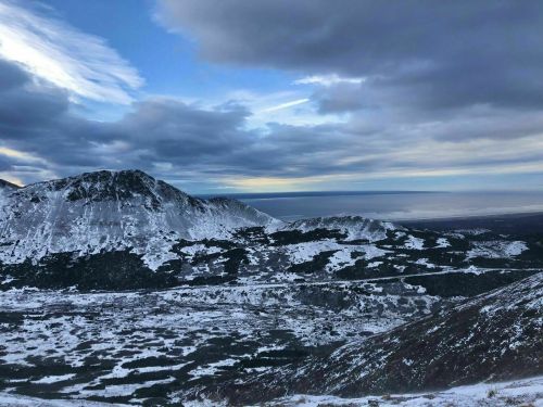 A panoramic view of snow-covered mountains under a cloudy sky, with a distant horizon and patches of blue.