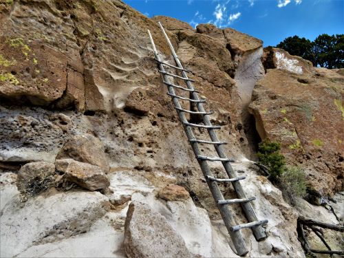 A wooden ladder leans against a rocky cliff, surrounded by textured stone and sparse vegetation under a blue sky.
