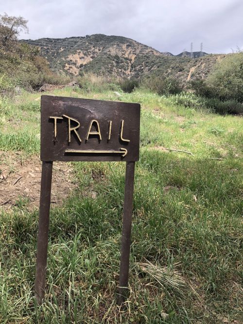 A wooden signpost labeled "TRAIL" points to a grassy path with hills in the background under a cloudy sky.