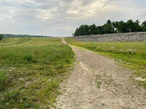 A gravel path leads through a grassy field, bordered by a stone wall and trees under a cloudy sky.
