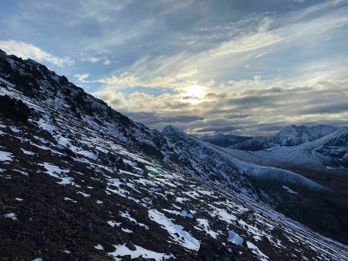 A snowy mountain landscape under a cloudy sky, with the sun peeking through the clouds in the background.