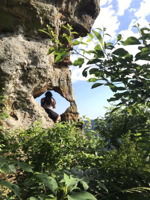 A person sits on a rock ledge, framed by greenery, gazing out at a scenic view under a blue sky.