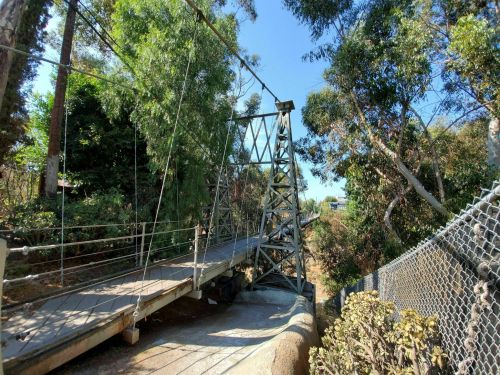 A suspension bridge surrounded by trees, spanning a dry creek bed under a clear blue sky.