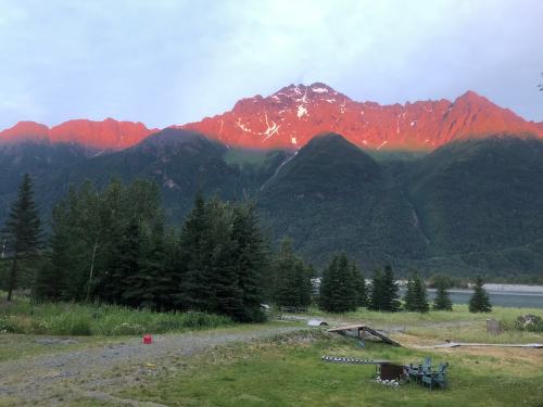 Sunset casts a warm glow on rugged mountains, surrounded by lush trees and a serene landscape.