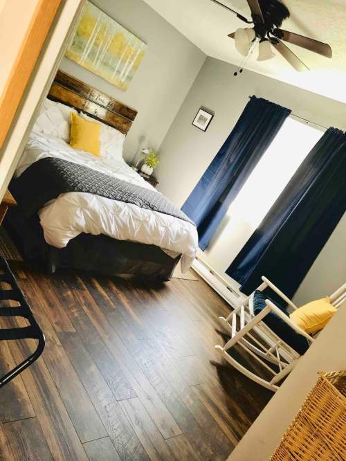 Cozy bedroom with a wooden bed, yellow accents, dark curtains, and a rocking chair on hardwood floors.