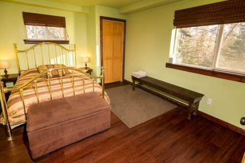 Cozy bedroom with a gold bed frame, green walls, wooden flooring, and a bench by the window. Natural light coming in.