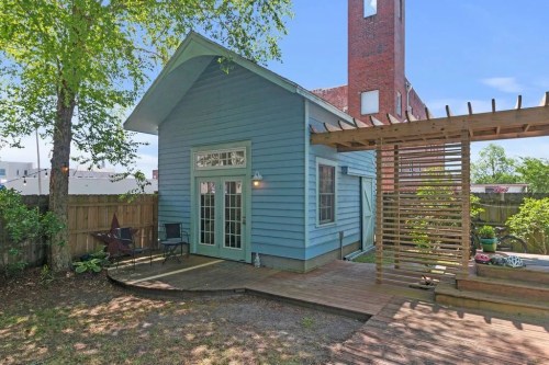 A charming blue house with a wooden deck and pergola in a backyard, surrounded by greenery and a wooden fence.