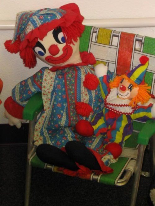 A large clown doll in a colorful outfit sits on a chair next to a smaller, brightly dressed clown plush toy.
