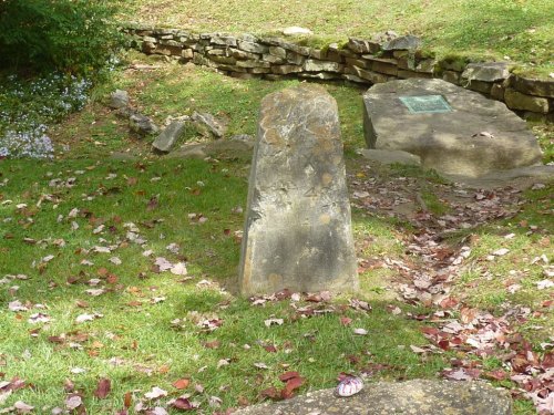 A weathered stone marker stands in a grassy area, surrounded by fallen leaves and a stone wall in the background.