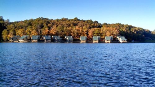 Row of cabins along a lake, surrounded by trees with autumn foliage under a clear blue sky.