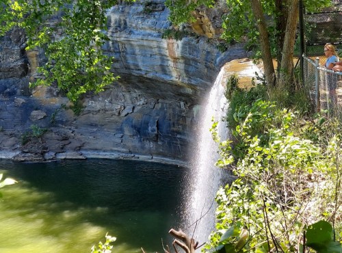 A waterfall cascades into a serene pool, surrounded by lush greenery and rocky cliffs. A person stands nearby, admiring the view.