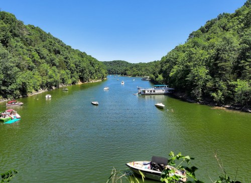 A serene river surrounded by lush green hills, with several boats floating on the calm water under a clear blue sky.