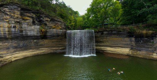 A serene waterfall cascades into a green pool, surrounded by rocky cliffs and lush trees. Two people swim nearby.
