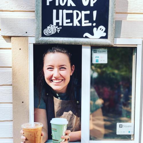 A smiling barista at a pickup window holds two coffee cups, with a sign above reading "Pick Up Here!"