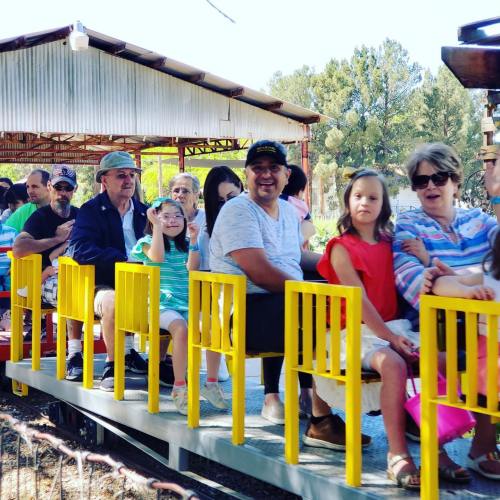 A group of people, including children and adults, sitting on a colorful train at an outdoor amusement area.