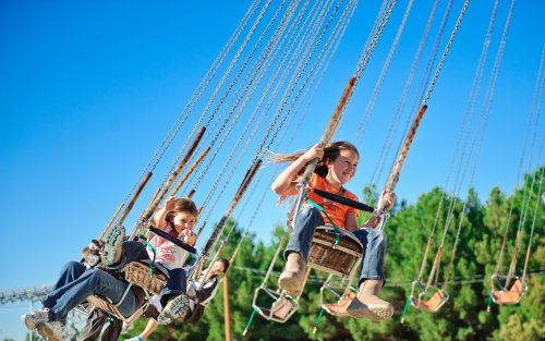 Two children enjoy a swing ride at an amusement park, smiling against a clear blue sky and green trees in the background.