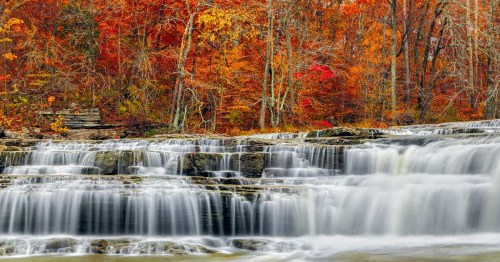 A serene waterfall cascades over rocks, surrounded by vibrant autumn foliage in shades of red, orange, and yellow.
